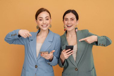 Sarı arka planda izole edilmiş cep telefonlarını kullanan ve işaret eden iki genç kadın.