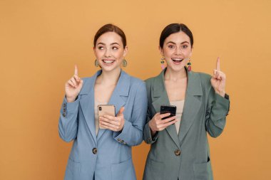 İki genç kadın cep telefonu kullanıyor ve sarı arka planda izole edilmiş olarak parmaklarını yukarı kaldırıyor.
