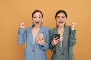 İki genç kadın cep telefonu kullanıyor ve kazanan hareketi sarı arka planda izole ediyor.