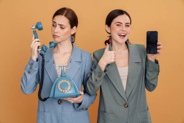Sarı arka planda izole edilmiş telefon ve akıllı telefonla poz veren iki genç kadın.