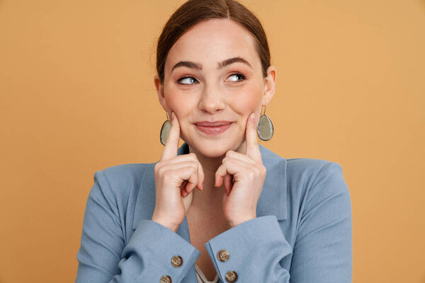 Young ginger woman smiling and pointing fingers at her cheeks isolated over yellow background