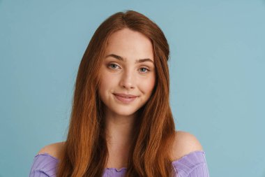Mavi duvarın arkasında dikilip kameraya bakan, uzun kızıl saçlı, güler yüzlü genç bir kadının portresini yakından çek.