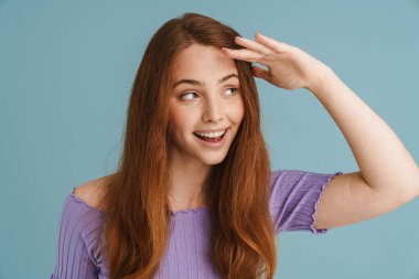 Genç kızıl kadın gülümseyip el sallarken mavi arka planda tek başına bir tarafa bakıyor.