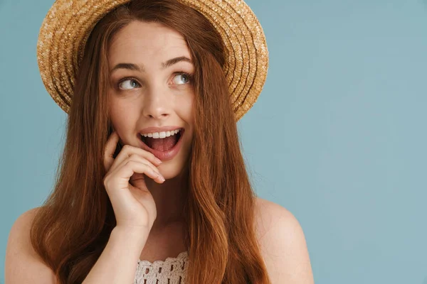 Hasır şapkalı genç kızıl kadın mavi arka planda mahsur kalmış kamerada şaşkınlığını ifade ediyor.