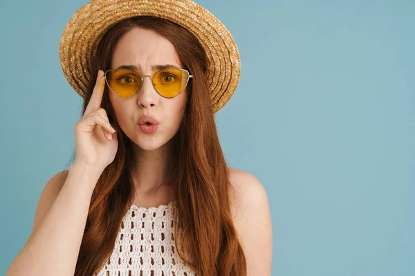 Güneş gözlüklü genç kızıl kadın mavi arka planda izole edilmiş kameraya şaşırdığını ifade ediyor.