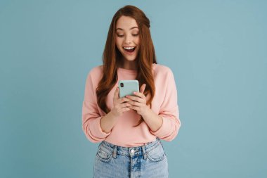 Mavi arka planda izole edilmiş, gülümseyen ve cep telefonu kullanan genç kızıl kadın.