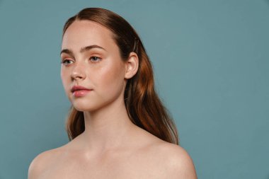 Mavi arka planda yarı çıplak kızıl kadın poz veriyor ve bir kenara bakıyor.