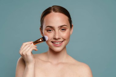 Yarı çıplak kızıl kadın gülümsüyor ve makyaj fırçası kullanıyor. Mavi arka planda izole edilmiş.