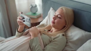 Pozitif sarışın kadın battaniyenin arkasına saklanıyor ve evde yatakta telefona bakıyor.
