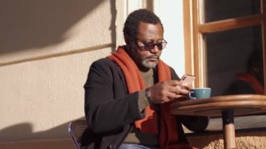Gözlüklü kendine güvenen Afrikalı adam telefonla bant yapıştırıyor ve açık kafede kahve içiyor.