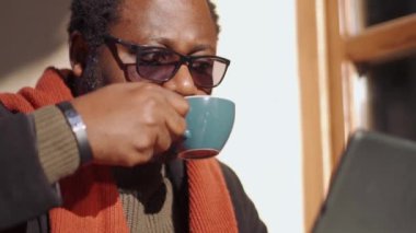 Gözlüklü dalgın Afrikalı adam tablete bakıyor ve açık kafede kahve içiyor.
