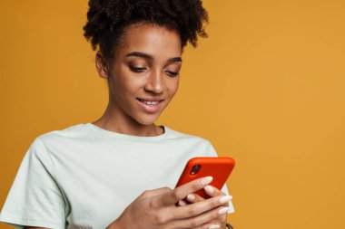 Young black woman smiling while using mobile phone isolated over yellow background