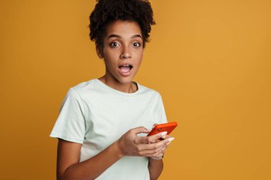 Young black woman expressing surprise while using mobile phone isolated over yellow background