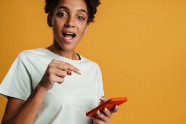 Young black woman using and pointing finger at mobile phone isolated over yellow background