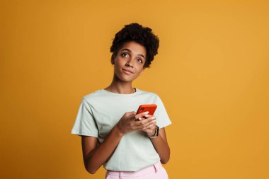 Young black woman smiling while using mobile phone isolated over yellow background