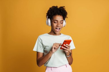 Young black woman listening music with headphones and cellphone isolated over yellow background