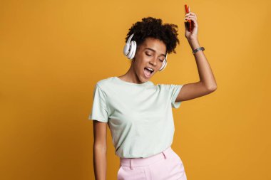 Young black woman listening music with headphones and singing isolated over yellow background