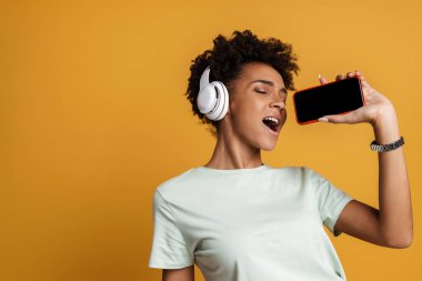 Young black woman listening music with headphones and singing isolated over yellow background