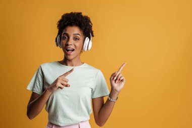 Young black woman with headphones smiling and pointing her fingers upward isolated over yellow background