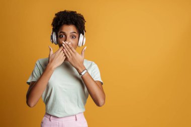 Young black woman using headphones and covering her face with arms isolated over yellow background