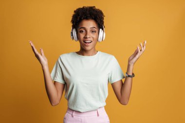 Young black woman with headphones smiling and throwing up her hands isolated over yellow background
