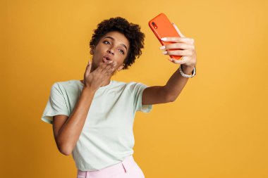 Young black woman smiling while using mobile phone isolated over yellow background