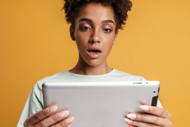 Young black woman expressing surprise while using tablet computer isolated over yellow background