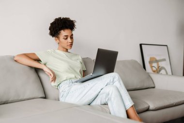 Young black woman working with laptop while sitting on couch at home