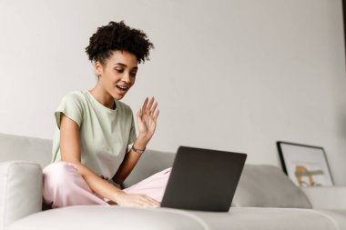 Young black woman gesturing and using laptop while sitting on couch at home