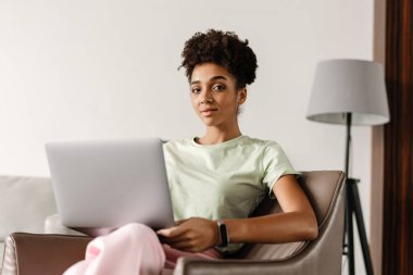 Young black woman working with laptop while sitting on armchair at home
