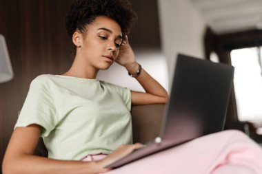 Young black woman working with laptop while sitting on armchair at home