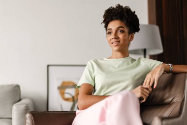 Young black woman smiling at camera while sitting on armchair at home