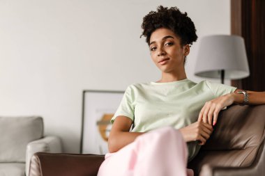 Young black woman looking at camera while sitting on armchair at home