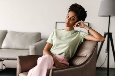 Young black woman looking at camera while sitting on armchair at home