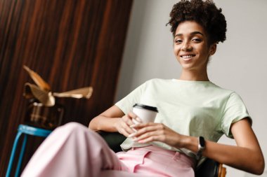 Young black woman smiling and drinking coffee while sitting on armchair at home