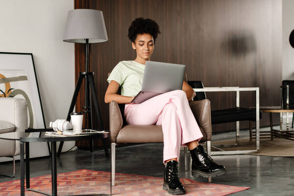 Young black woman working with laptop while sitting on armchair at home