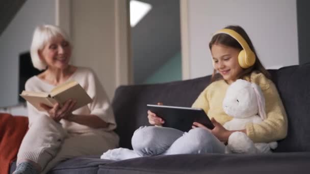 Positive Girl Headphones Looking Tablet Talking Her Grandmother Reading Book — Vídeo de Stock