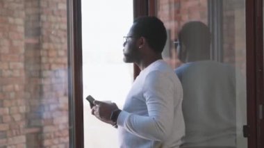 Konsantre olmuş Afrikalı adam telefonla mesaj atıyor ve pencerenin yanında kahve içiyor.
