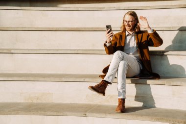 White ginger man gesturing and using cellphone while sitting on stairs outdoors