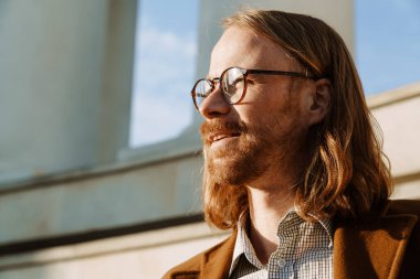 White ginger man wearing jacket and eyeglasses looking aside outdoors
