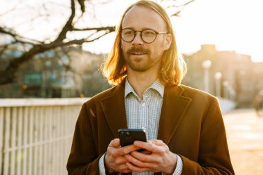 White ginger man in eyeglasses smiling while using mobile phone at city street