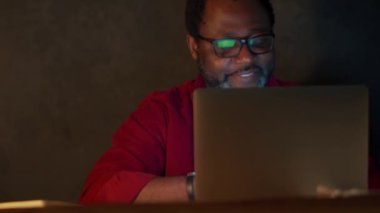 Cheerful African man working on laptop in office at night