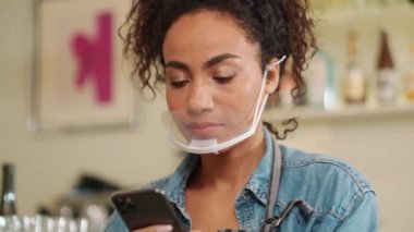 Laughing African woman barista in protective mask texting by phone in cafe