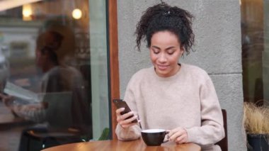 Pensive African woman drinking coffee and typing by mobile outdoors