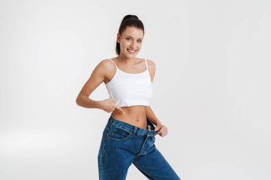 European woman pointing finger at her waist while posing in big jeans isolated over white background