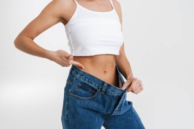 European woman pointing finger at her waist while posing in big jeans isolated over white background
