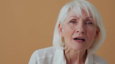 Beyaz gömlekli, bej renkli stüdyoda kameraya bakan mutlu olgun bir kadın.