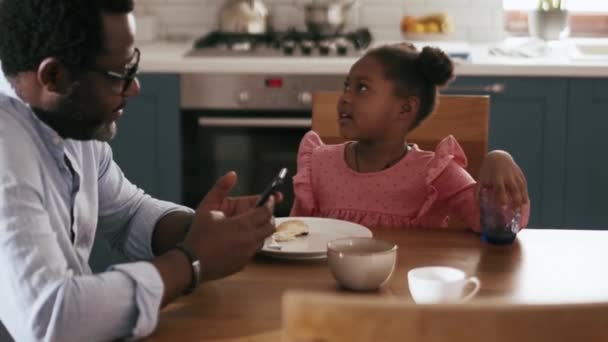 アフリカの母親クリア料理をオフにし 彼女の娘と話をしながら 父親は台所で電話を見て — ストック動画