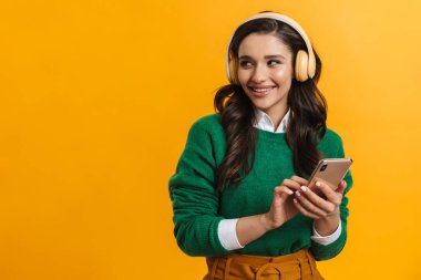 Mutlu gülümseyen sıradan bir kadının portresi sarı arka planda durup elinde cep telefonu olan kulaklıkla müzik dinliyor.