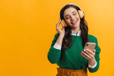 Mutlu gülümseyen sıradan bir kadının portresi sarı arka planda durup elinde cep telefonu olan kulaklıkla müzik dinliyor.